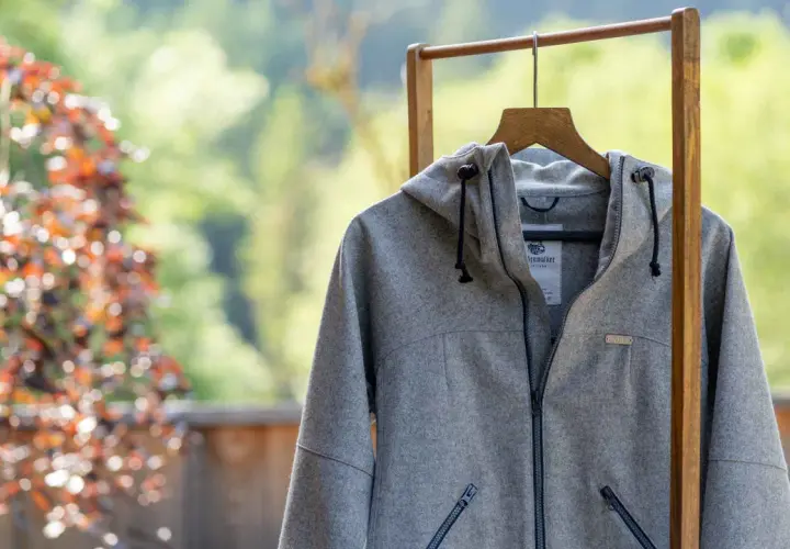 Graue Lodenjacke mit Kapuze, Reißverschluss und Taschen, aufgehängt an einem Holzständer vor blühendem Baum und grüner Natur.