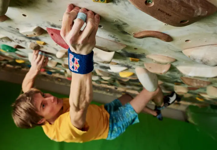 Alex Megos klettert konzentriert an einer Boulderwand. Seine Hand greift fest einen Griff, während sein Blick nach oben gerichtet ist.