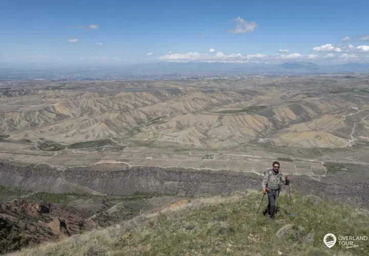 Ein Mann mit Rucksack und Wanderstöcken genießt die weite Aussicht bei seinem Armenien Wandern. Die Landschaft ist geprägt von trockenen Hügeln und einer fernen Stadt.