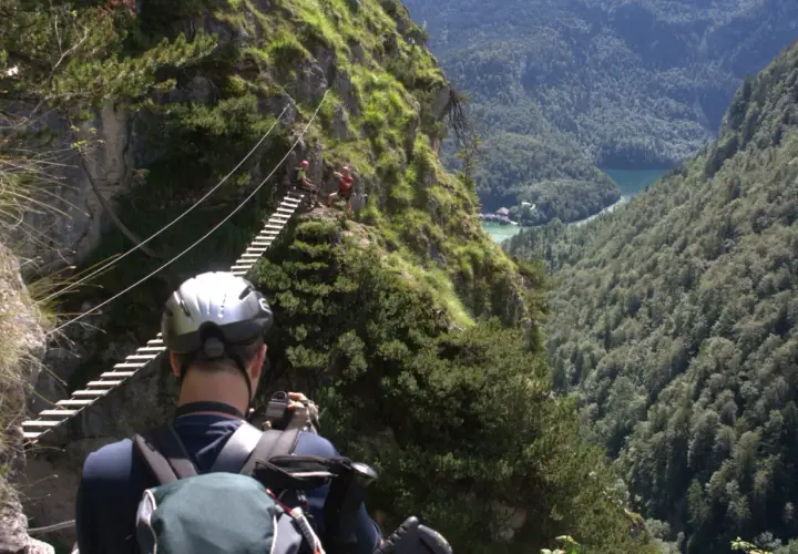 Ein Mann mit Helm und Rucksack fotografiert einen Klettersteig in Berchtesgaden. Eine Hängebrücke führt über eine Schlucht mit Blick auf einen See.
