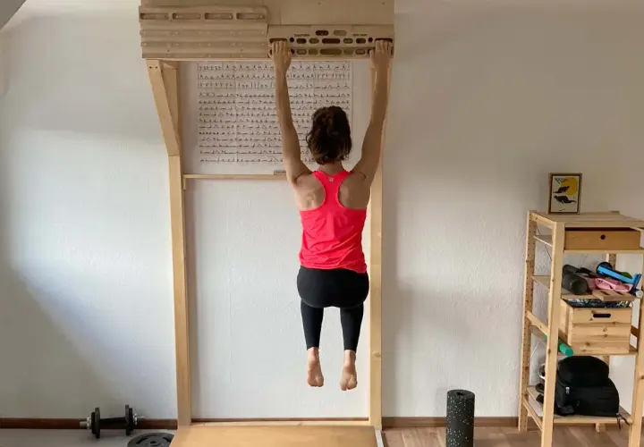 Frau hängt an einem Hangboard für intensives **Hangboard Training**. Trainingsplan hängt an der Wand.
