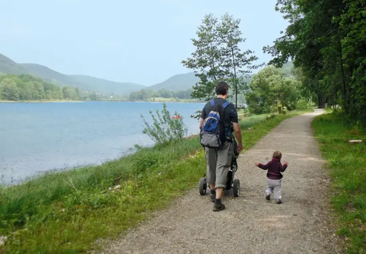 Vater schiebt Kinderwagen am Happurger Stausee entlang, während das Kind vorausläuft. Herrliche Kulisse für eine Wanderung.
