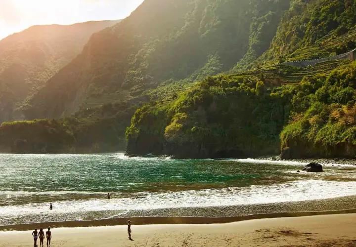 Sonne beleuchtet den malerischen **Madeira schwarzer Strand** mit üppig grünen Bergen im Hintergrund. Wellen brechen sanft am Ufer.