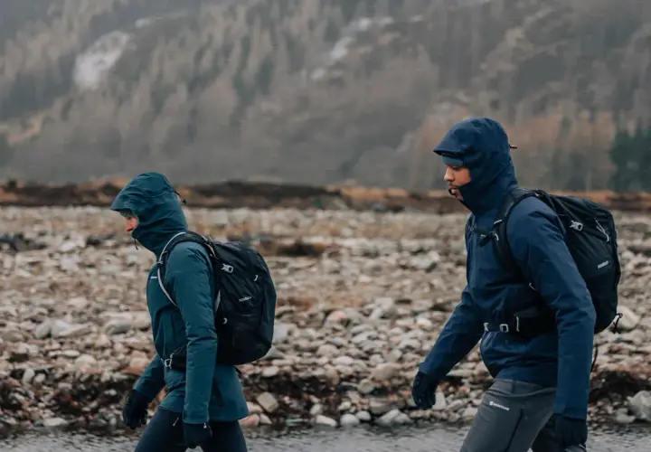 Zwei Wanderer mit Rucksäcken gehen durch eine steinige Landschaft in Schottland. Das Wetter ist regnerisch, die Kapuzen sind hochgezogen.