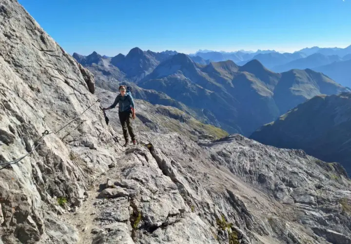 Frau auf dem **Heilbronner Weg wandern**, gesichert an einem Stahlseil, mit atemberaubendem Bergpanorama im Hintergrund.