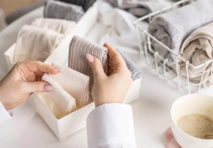 Hände sortieren gefaltete Kleidung, darunter auch warme **Wandersocken**, in einem Behälter. Daneben ein Korb mit weiteren Textilien und eine Kaffeetasse.