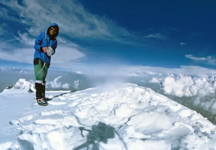 Reinhard Messner steht auf einem schneebedeckten Gipfel, umgeben von Wolken und blauem Himmel.