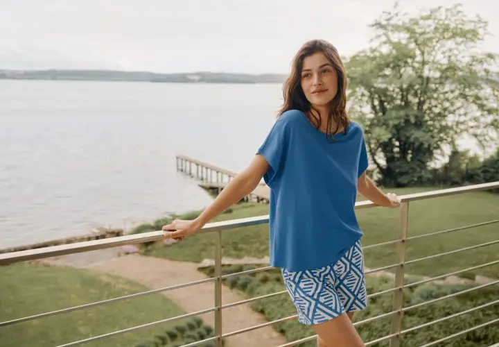 Frau auf Balkon mit Blick auf See, trägt blaues Oberteil aus modal stoff und gemusterte Shorts.