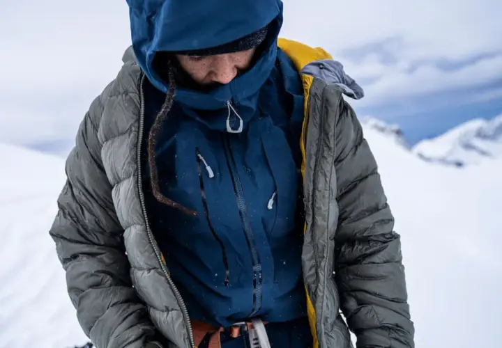 Person in Daunenjacke im Schnee. Die Jacke ist grau, darunter eine blaue Schicht. Perfekt für das Daunenjacke waschen nach einer Bergtour.