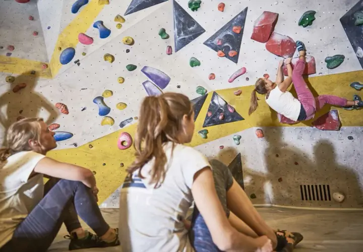 Zwei Mädchen schauen zu, wie eine dritte Person an der Kletterwand hochklettert. Sie tragen bequeme Kleidung, wie man sie zum Bouldern anzieht: Leggings und T-Shirts.