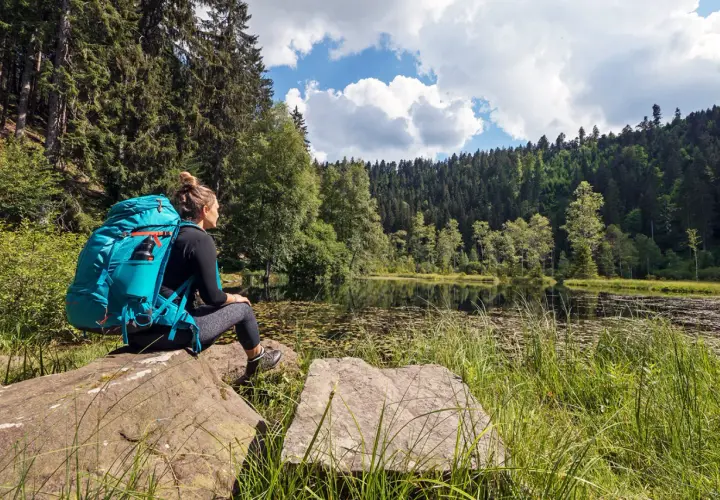 Frau mit Rucksack sitzt an einem See, umgeben von Wald. Ein perfekter Moment auf einer der vielen **weitwanderwege europa**.