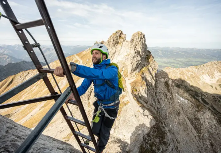 Mann klettert auf einem Leiternabschnitt des Oberstdorf Klettersteigs. Die Aussicht auf die Alpen ist atemberaubend.