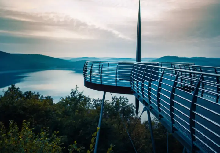 Moderne Aussichtsplattform über einem See, ideal für das Wandern in NRW. Sanfte Hügel und ein dramatischer Himmel im Hintergrund.