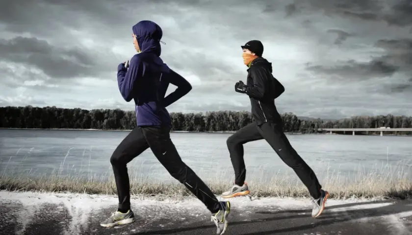 Zwei Läuferinnen in warmer Winterkleidung joggen am Fluss entlang. Die eine trägt einen blauen Hoodie und Hijab, die andere eine schwarze Jacke und orangefarbenen Schal.