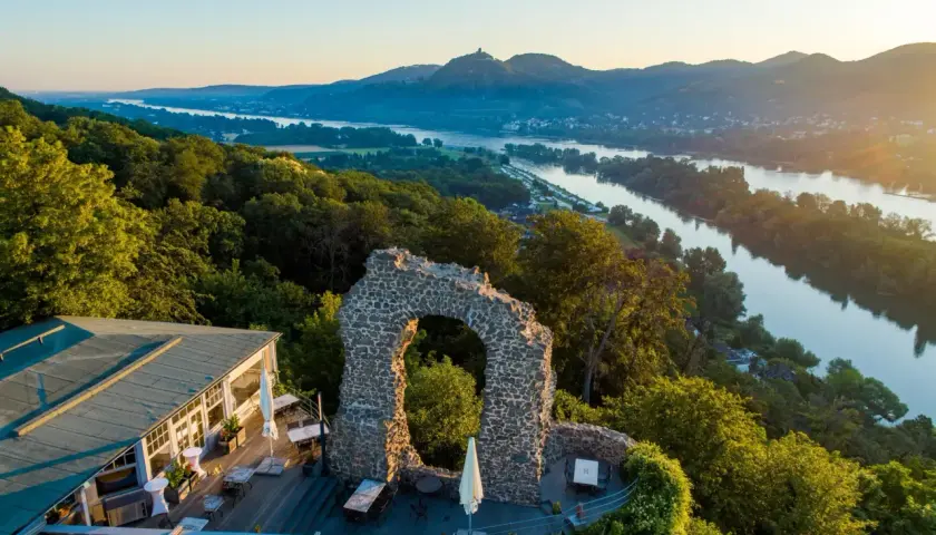 Der Rolandbogen thront über dem Rhein. Ein Restaurant mit Terrasse lädt zum Verweilen ein, während im Hintergrund Berge und ein Fluss im Abendlicht glänzen.