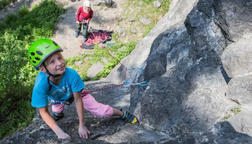 Ein Mädchen mit Helm klettert an einem Felsen, während eine andere Person sichert. Ein tolles Erlebnis beim Klettern mit Kindern.