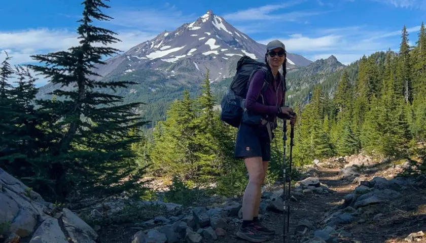 Frau mit Rucksack und Wanderstöcken auf einem steinigen Wanderweg in den USA, im Hintergrund ein schneebedeckter Berg und Nadelbäume.
