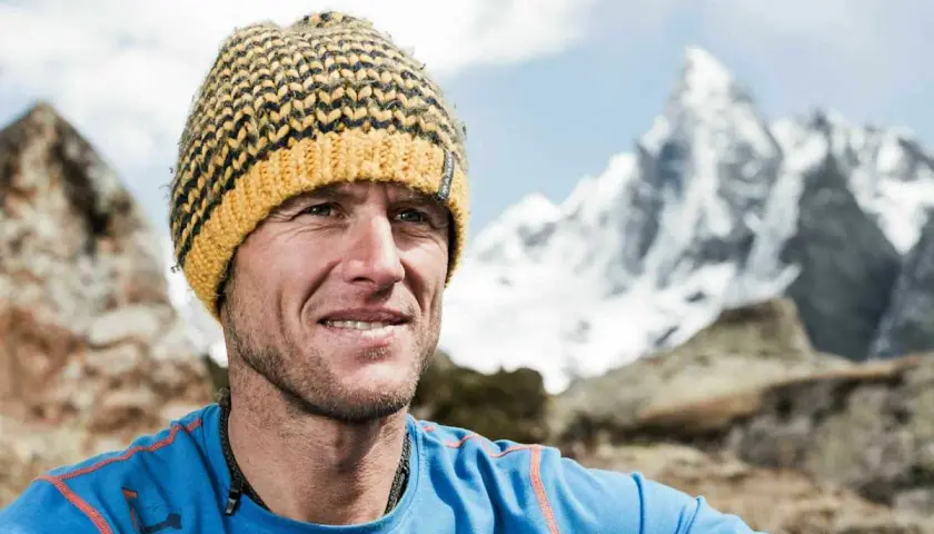 Stephan Siegrist, ein Bergsteiger, lächelt in die Kamera, eine gestreifte Mütze auf dem Kopf, vor einer schneebedeckten Bergkulisse.