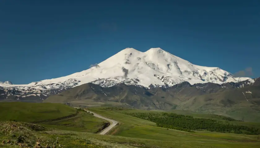 Der majestätische Elbrus Berg, bedeckt mit Schnee, ragt über grüne Hügel und eine kurvige Straße auf.