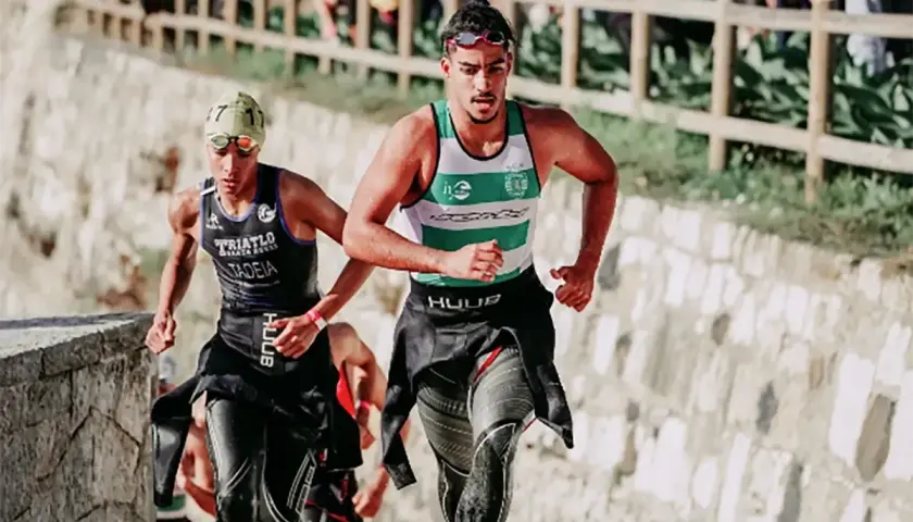 Zwei Athleten laufen bergauf, die Triathlon-Reihenfolge im Blick. Der vordere trägt ein grünes und weißes Shirt, der hintere ein schwarzes mit der Aufschrift "TRIATLO".