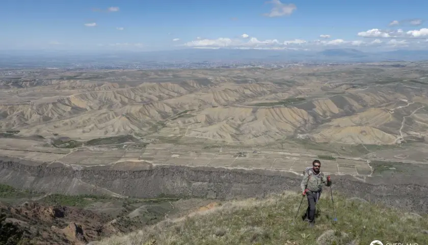Ein Mann mit Rucksack und Wanderstöcken genießt die weite Aussicht bei seinem Armenien Wandern. Die Landschaft ist geprägt von trockenen Hügeln und einer fernen Stadt.