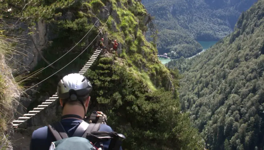 Ein Mann mit Helm und Rucksack fotografiert einen Klettersteig in Berchtesgaden. Eine Hängebrücke führt über eine Schlucht mit Blick auf einen See.