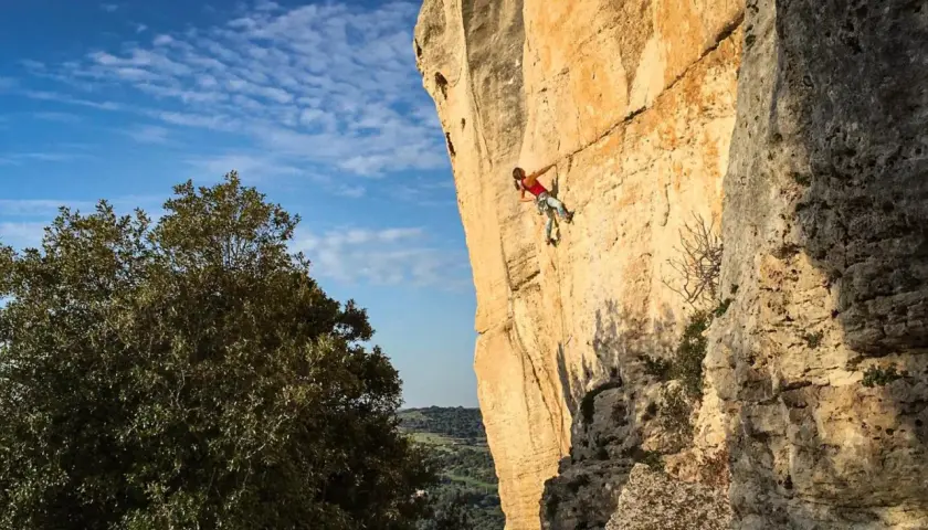 Eine Frau klettert an einer sonnenbeschienenen Felswand in Sardinien. Der blaue Himmel mit Wolken und grüne Bäume bilden den Hintergrund.