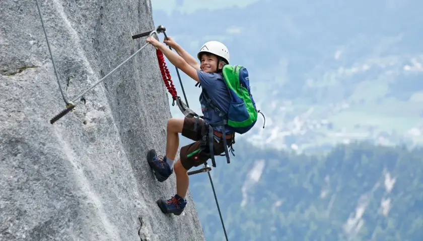 Ein Junge mit Helm und Rucksack klettert einen Felsgrat. Ein spannendes Abenteuer für klettersteig kinder.