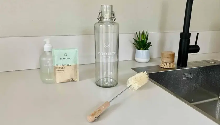 Alles für die Edelstahl Trinkflasche reinigen: Spülmittel, Pulver, Flaschenbürste und ein kleiner Topf mit Pflanze.