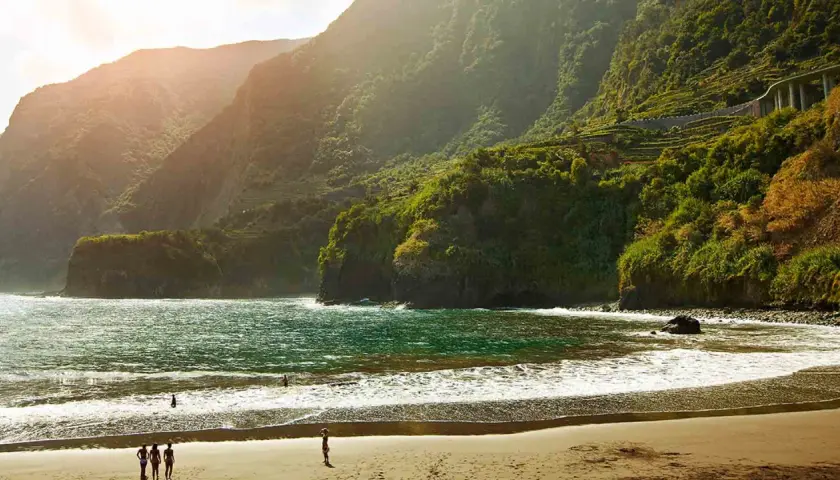 Sonne beleuchtet den malerischen **Madeira schwarzer Strand** mit üppig grünen Bergen im Hintergrund. Wellen brechen sanft am Ufer.