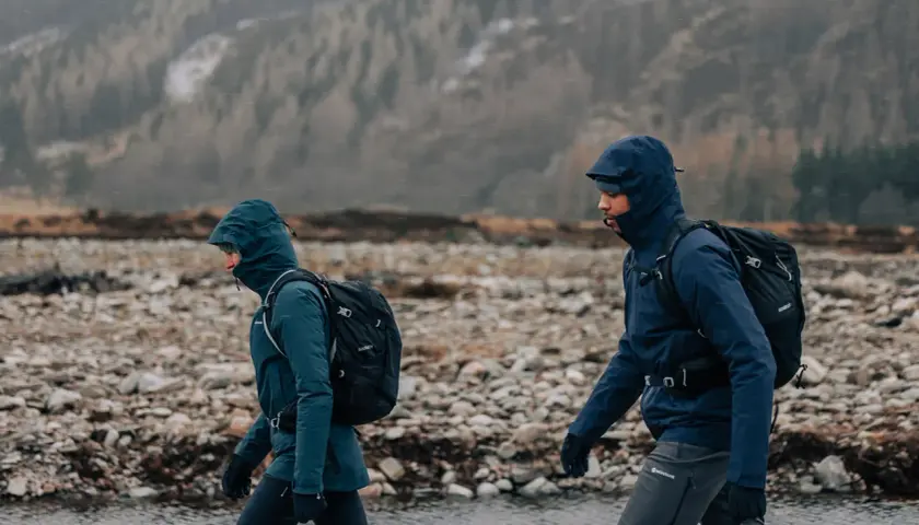 Zwei Wanderer mit Rucksäcken gehen durch eine steinige Landschaft in Schottland. Das Wetter ist regnerisch, die Kapuzen sind hochgezogen.