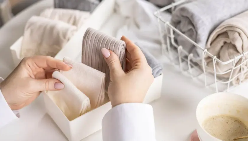 Hände sortieren gefaltete Kleidung, darunter auch warme **Wandersocken**, in einem Behälter. Daneben ein Korb mit weiteren Textilien und eine Kaffeetasse.