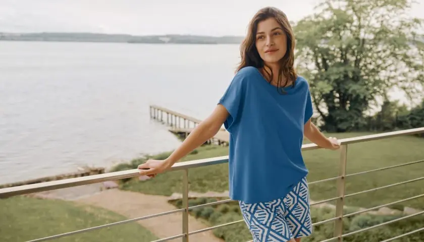 Frau auf Balkon mit Blick auf See, trägt blaues Oberteil aus modal stoff und gemusterte Shorts.