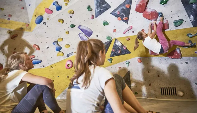 Zwei Mädchen schauen zu, wie eine dritte Person an der Kletterwand hochklettert. Sie tragen bequeme Kleidung, wie man sie zum Bouldern anzieht: Leggings und T-Shirts.