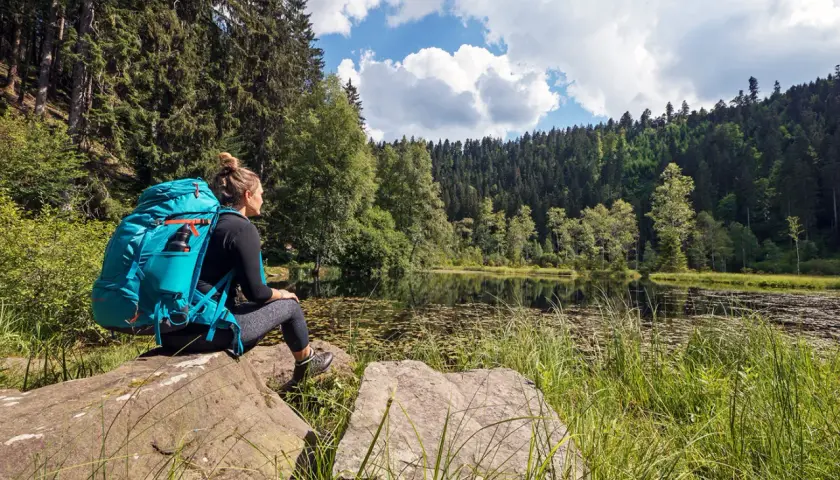 Frau mit Rucksack sitzt an einem See, umgeben von Wald. Ein perfekter Moment auf einer der vielen **weitwanderwege europa**.