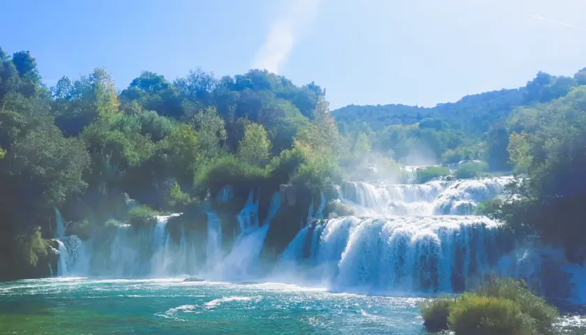 Ein majestätischer Krk Wasserfall stürzt über mehrere Stufen in ein türkisfarbenes Becken. Üppige grüne Bäume säumen die Klippen und der Himmel ist strahlend blau.