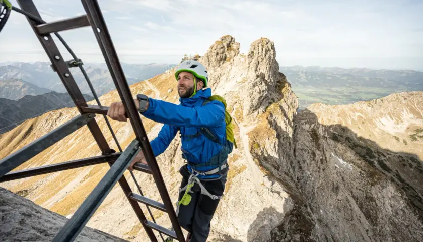 Mann klettert auf einem Leiternabschnitt des Oberstdorf Klettersteigs. Die Aussicht auf die Alpen ist atemberaubend.