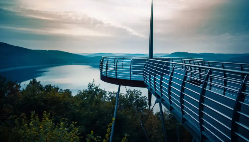 Moderne Aussichtsplattform über einem See, ideal für das Wandern in NRW. Sanfte Hügel und ein dramatischer Himmel im Hintergrund.