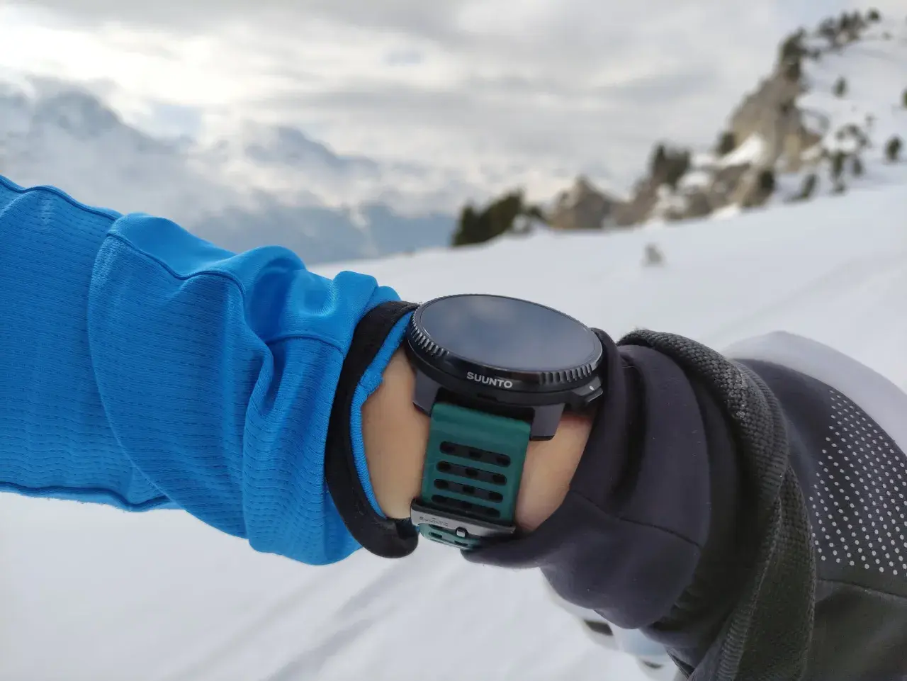 Suunto Vertical Test: Arm in blauer Jacke mit grüner Suunto-Uhr im Schnee, Bergkulisse.