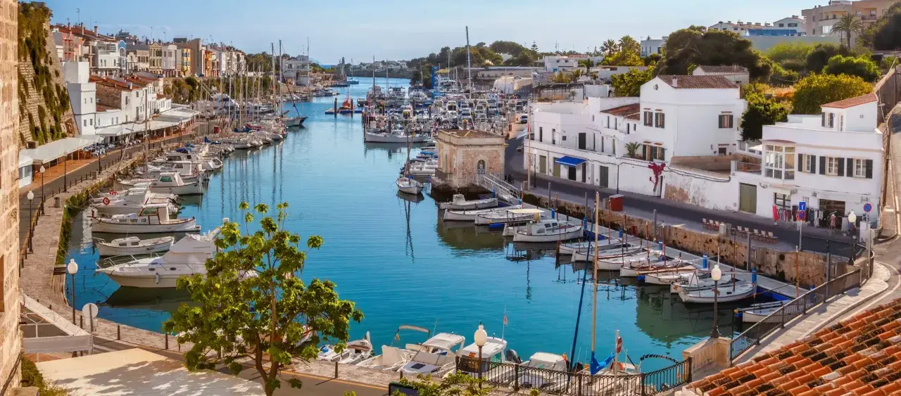 Hafen von Ciutadella, Menorca. Boote im türkisfarbenen Wasser, weiße Häuser und bunte Fassaden. Ein malerischer Blick auf die Menorca Highlights.