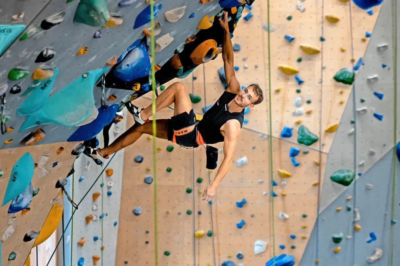 Yannick Flohé klettert an einer Boulderwand, hält sich an einem bunten Griff fest und blickt lächelnd in die Kamera.