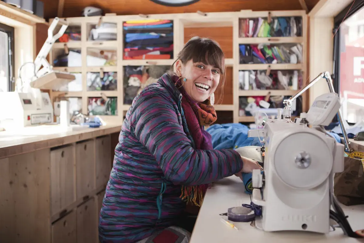 Frau lacht fröhlich an ihrer Nähmaschine, bereit für die Patagonia Reparatur. Regale voller bunter Outdoor-Kleidung im Hintergrund.