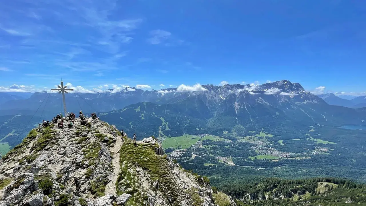 Gipfelkreuz auf dem Kramer­spitz, umgeben von Wanderern. Im Hintergrund das Alpenpanorama und ein Tal mit Häusern.