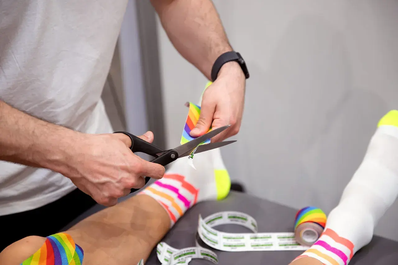 Hände schneiden buntes Kinesio-Tape zu. Die Frage "kinesio tape wann entfernen" wird hier visuell beantwortet: wenn es zugeschnitten wird, ist es bereit zur Anwendung.