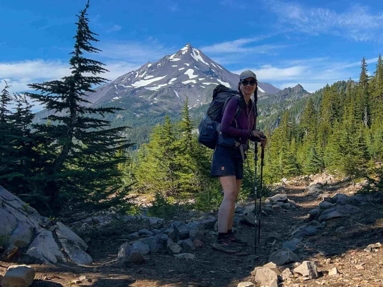 Frau mit Rucksack und Wanderstöcken auf einem steinigen Wanderweg in den USA, im Hintergrund ein schneebedeckter Berg und Nadelbäume.