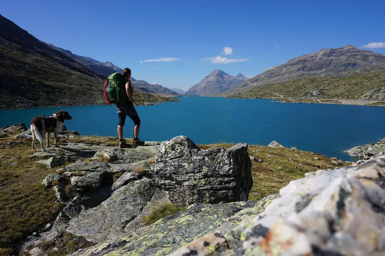 Wanderer mit Rucksack und Hund genießen den Ausblick auf einen türkisfarbenen See in den Alpen. Perfekt für mehrtageswanderungen.