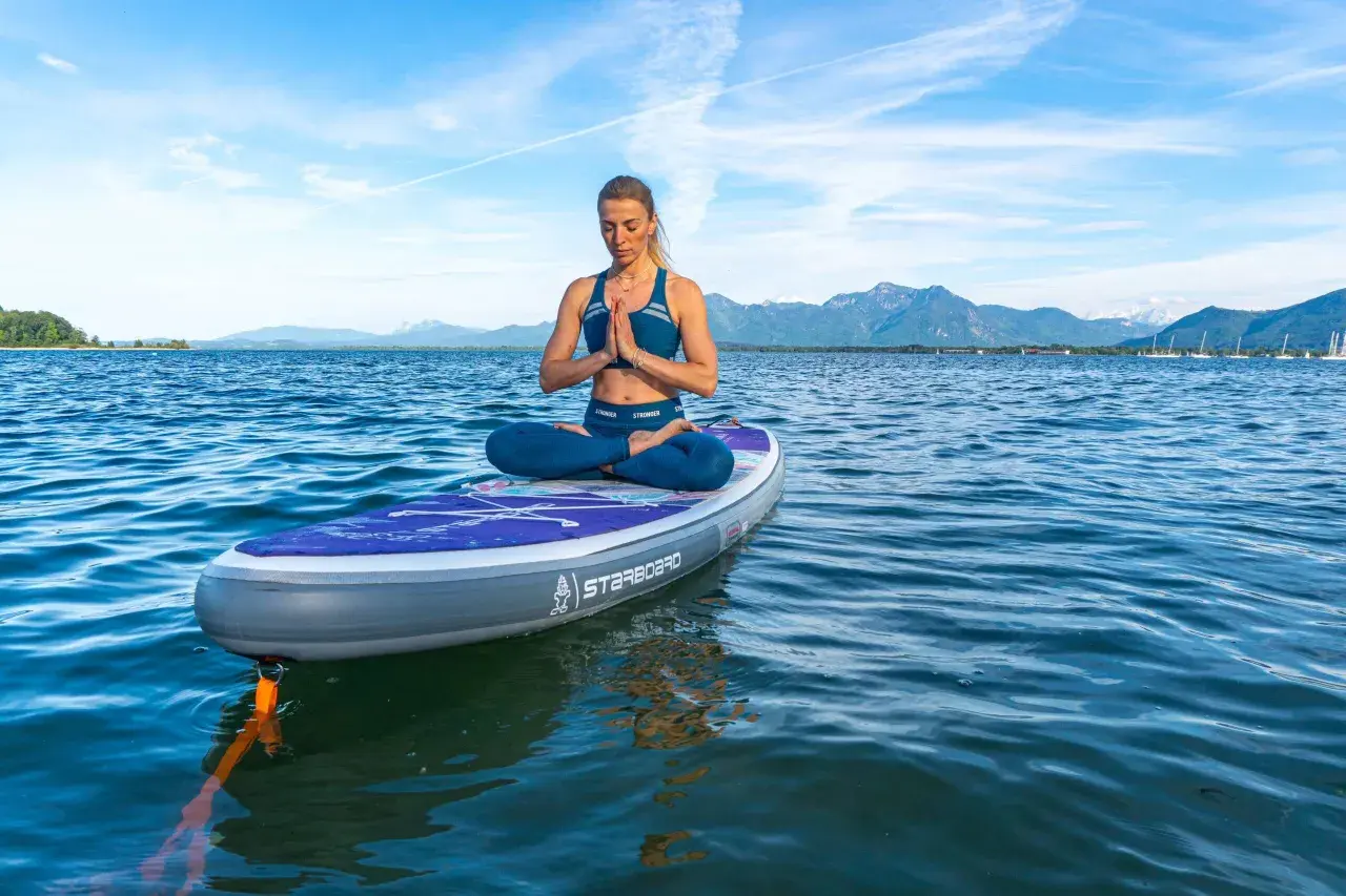 Frau praktiziert SUP Yoga auf einem Brett im See, mit Bergen im Hintergrund.