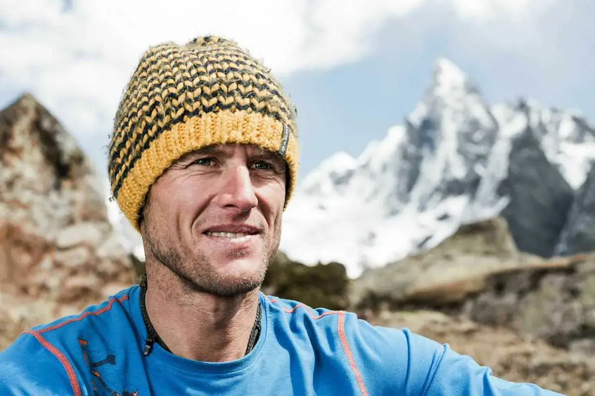 Stephan Siegrist, ein Bergsteiger, lächelt in die Kamera, eine gestreifte Mütze auf dem Kopf, vor einer schneebedeckten Bergkulisse.