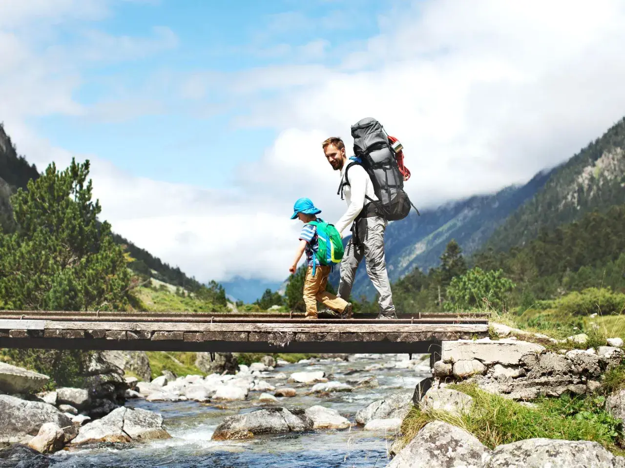 Vater und Sohn überqueren eine Brücke beim Wandern mit Kleinkind. Berge und ein Fluss im Hintergrund.