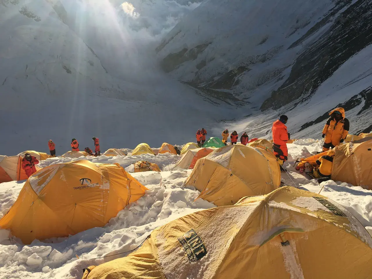 Bergsteiger in orangefarbenen Anzügen bereiten sich auf den Aufstieg vom Mount Everest Camp 4 vor. Gelbe Zelte sind im Schnee verstreut.